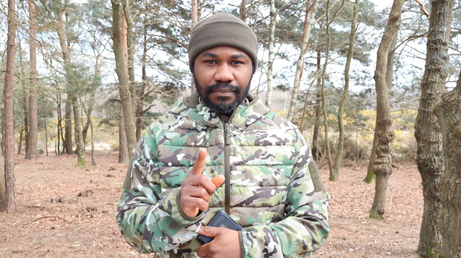 Raphael Rabbi, Overseer of Invisible War Army, standing in a forest in camo attire, holding a Bible and raising one finger, with scripture and ministry information overlaid on the image